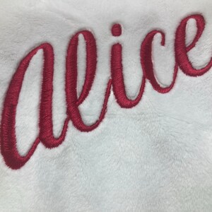 May include: White plush fabric with the embroidered name "Alice" in a cursive, deep red font. The embroidery has a textured appearance, and the fabric has a soft, fluffy texture. The name is centered and takes up a large portion of the image.