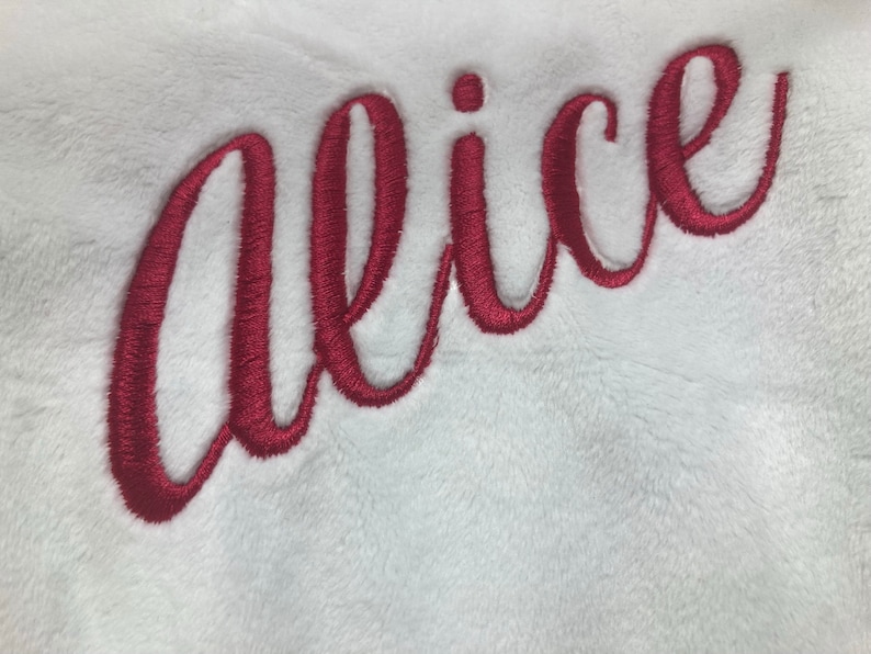 May include: White plush fabric with the embroidered name "Alice" in a cursive, deep red font. The embroidery has a textured appearance, and the fabric has a soft, fluffy texture. The name is centered and takes up a large portion of the image.