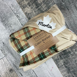 May include: A beige and green plaid cabin blanket with a cream-colored fleece lining. The blanket is folded and tied with a white ribbon. The blanket has a white maple leaf with the name "Martin" embroidered on it. The label reads "Kawartha Pillow Co. Cabin Blanket".
