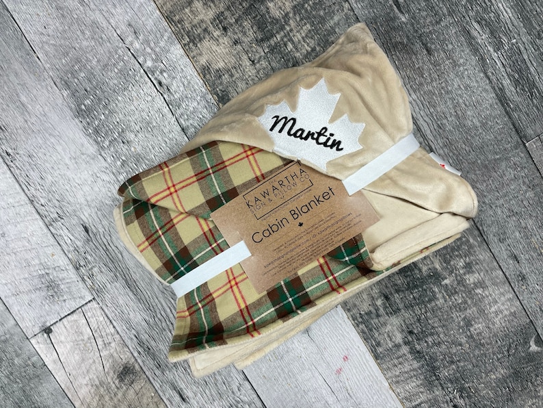 May include: A beige and green plaid cabin blanket with a cream-colored fleece lining. The blanket is folded and tied with a white ribbon. The blanket has a white maple leaf with the name "Martin" embroidered on it. The label reads "Kawartha Pillow Co. Cabin Blanket".
