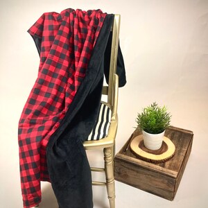 May include: A red and black buffalo plaid throw blanket with a black fleece backing. The blanket is draped over a wooden chair with a black and white striped cushion.