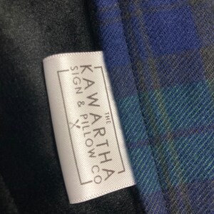 May include: Close-up of a white tag with the text "KAWARTHA SIGN & PILLOW CO" in a rectangular frame. The tag is next to a blue, green, and gray plaid fabric and a black fabric.