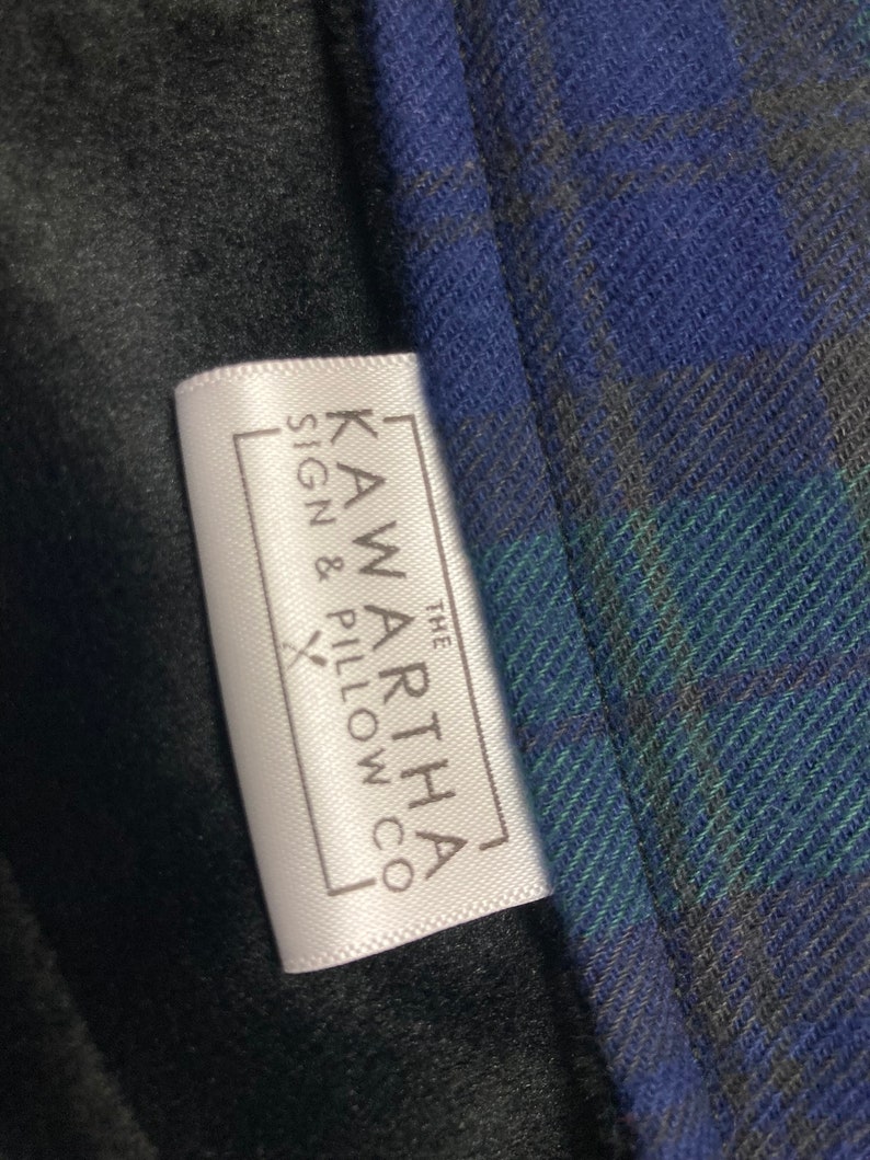 May include: Close-up of a white tag with the text "KAWARTHA SIGN & PILLOW CO" in a rectangular frame. The tag is next to a blue, green, and gray plaid fabric and a black fabric.
