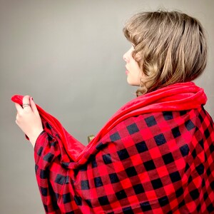 May include: A red and black plaid blanket with a soft, red fleece lining. The blanket is draped over a person's shoulders.