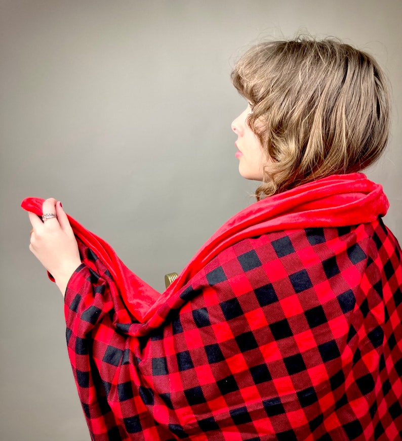 May include: A red and black plaid blanket with a soft, red fleece lining. The blanket is draped over a person's shoulders.