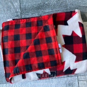 May include: Folded blanket featuring a red and black buffalo plaid pattern on one side and a white background with red and black maple leaf designs on the other. A small tag with a Canadian flag is visible.
