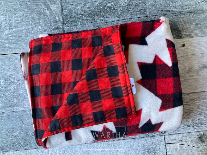 May include: Folded blanket featuring a red and black buffalo plaid pattern on one side and a white background with red and black maple leaf designs on the other. A small tag with a Canadian flag is visible.