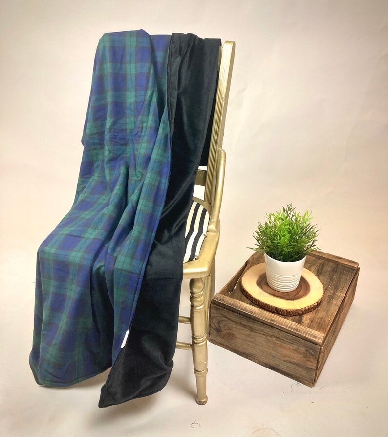 May include: A blue and green plaid blanket draped over a gold chair, with a black blanket folded over it. A small plant in a white pot sits on a wooden tray.