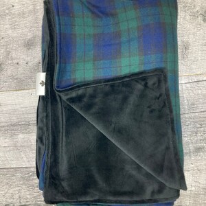 May include: Folded blanket with a plaid pattern in shades of blue and green, with a black plush underside. The blanket is folded neatly, showcasing the contrasting textures and colors. A small white tag with a leaf logo is visible.