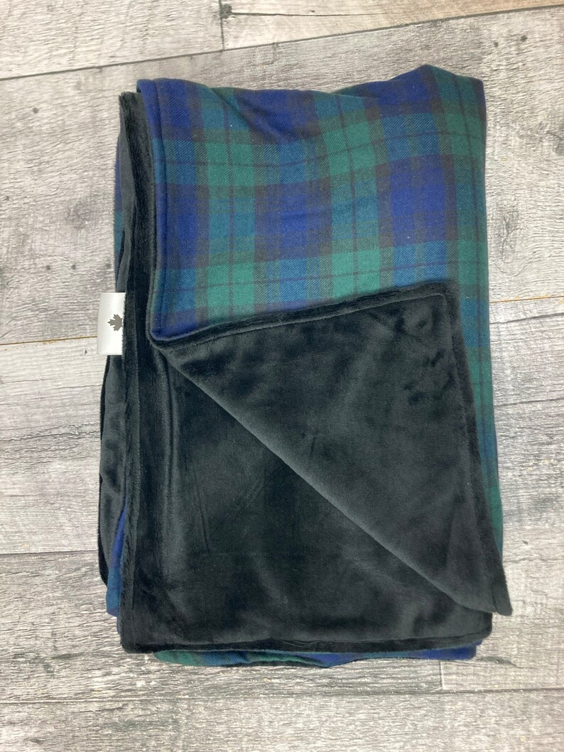 May include: Folded blanket with a plaid pattern in shades of blue and green, with a black plush underside. The blanket is folded neatly, showcasing the contrasting textures and colors. A small white tag with a leaf logo is visible.