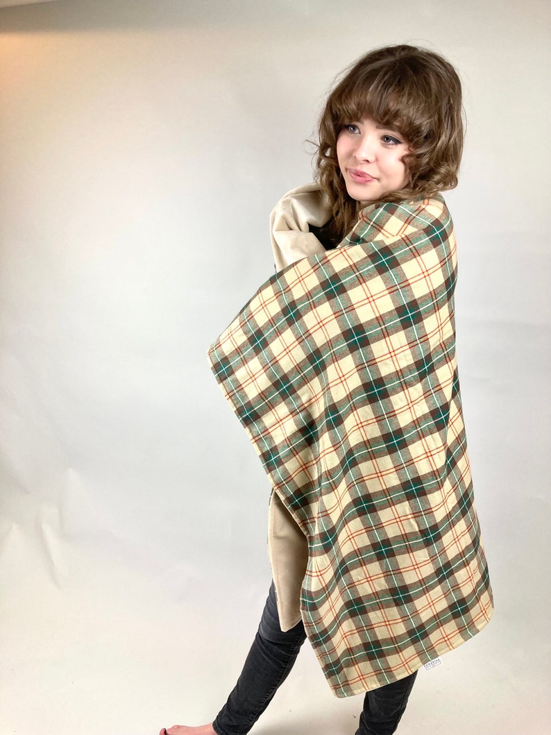 May include: A person wearing black jeans and a plaid blanket with brown, green, and white squares. The blanket is folded over the person's shoulders and is being held in their arms.