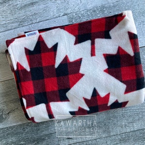 May include: Folded fleece blanket featuring a red and black plaid pattern with large white maple leaf designs. The blanket has a soft texture and is folded neatly. The text "KAWARTHA SIGN & PILLOW CO" is visible on a tag.