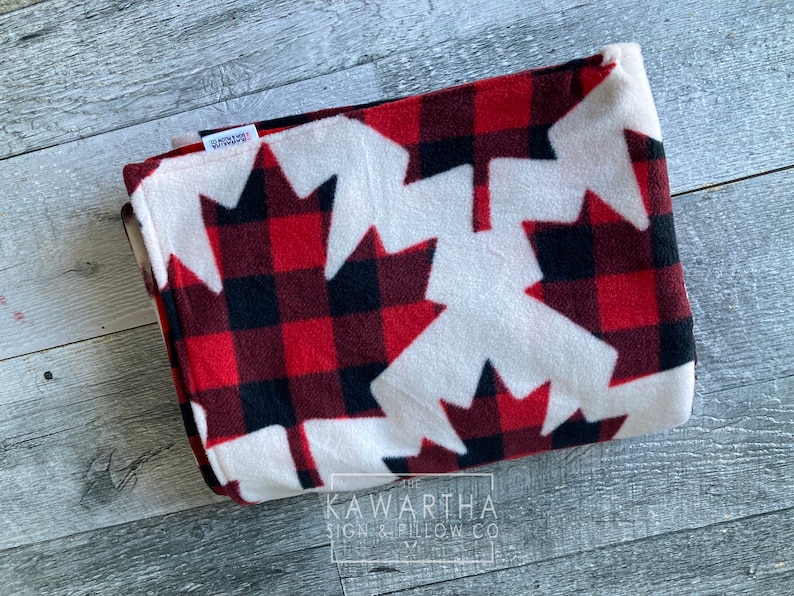 May include: Folded fleece blanket featuring a red and black plaid pattern with large white maple leaf designs. The blanket has a soft texture and is folded neatly. The text "KAWARTHA SIGN & PILLOW CO" is visible on a tag.