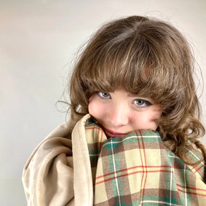 May include: A young person with brown hair is wrapped in a plaid blanket with green, brown, and yellow colors. The blanket is folded over the person's face, and they are looking at the camera with a smile.