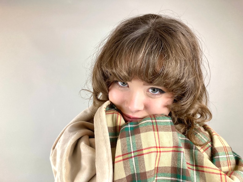May include: A young person with brown hair is wrapped in a plaid blanket with green, brown, and yellow colors. The blanket is folded over the person's face, and they are looking at the camera with a smile.