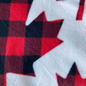 May include: A close-up of a soft, white fleece fabric with a repeating pattern of red and black plaid maple leaf shapes. The fabric has a cozy, textured appearance, perfect for blankets or apparel.