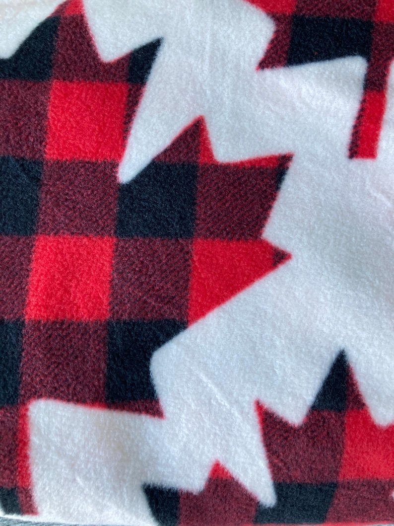 May include: A close-up of a soft, white fleece fabric with a repeating pattern of red and black plaid maple leaf shapes. The fabric has a cozy, textured appearance, perfect for blankets or apparel.