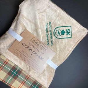 May include: A beige and green plaid cabin blanket with a white label that reads "The Kawartha Sign & Pillow Co. Cabin Blanket" and a green embroidered logo that says "Building Better Lives Through Swimming SK".