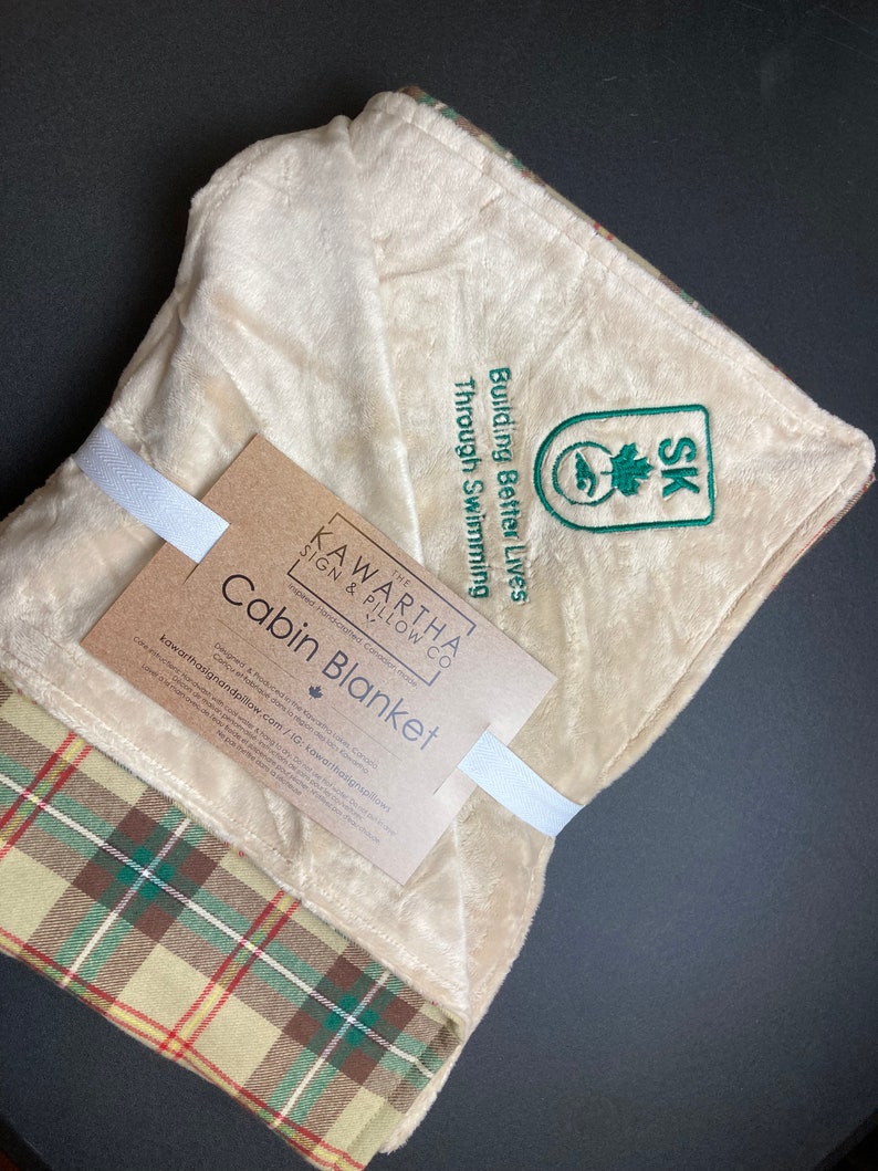 May include: A beige and green plaid cabin blanket with a white label that reads "The Kawartha Sign & Pillow Co. Cabin Blanket" and a green embroidered logo that says "Building Better Lives Through Swimming SK".