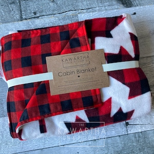 May include: Folded cabin blanket with a red and black buffalo plaid pattern on one side and a white background with red maple leaf designs on the other. The blanket is tied with a cream-colored ribbon and has a product tag.