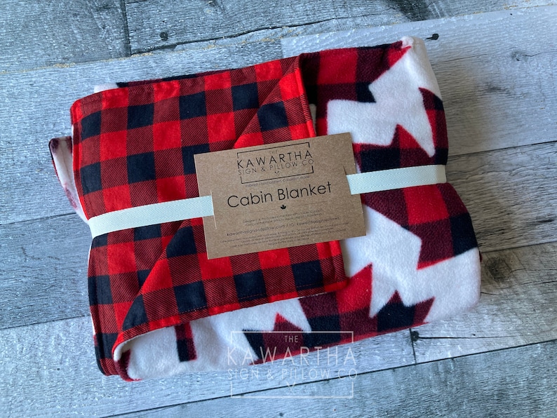 May include: Folded cabin blanket with a red and black buffalo plaid pattern on one side and a white background with red maple leaf designs on the other. The blanket is tied with a cream-colored ribbon and has a product tag.