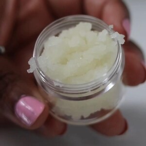 May include: A small, clear plastic jar filled with a light yellow, granular substance. The jar is held by a person with pink nail polish. The substance appears to be a cosmetic product, possibly a scrub or balm.