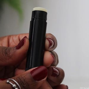 May include: A black lip balm tube with a white, rounded top is held between two hands with dark red nail polish. The lip balm is in a vertical position, and the background is blurred.