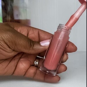 May include: A clear lip gloss tube filled with a shimmery, pink-toned gloss. The applicator is partially inserted, showing the product's texture. The gloss is held in a hand with a light pink manicure.