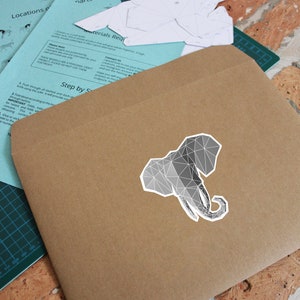 May include: A brown paper envelope with a geometric gray elephant sticker on the front. The envelope has instructions for a paper craft project printed on it.