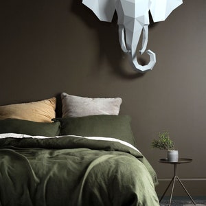 May include: A green linen duvet cover with a white stripe and a mustard yellow sheet underneath. The bed is styled with two pillows, one mustard yellow and one gray. A white origami elephant head is hanging on the wall above the bed.