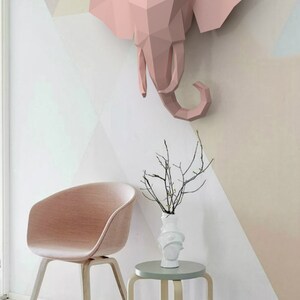 May include: A pink origami elephant head sculpture mounted on a white wall with a pink and white geometric pattern. A pink chair and a small wooden stool with a light green top are in the foreground.
