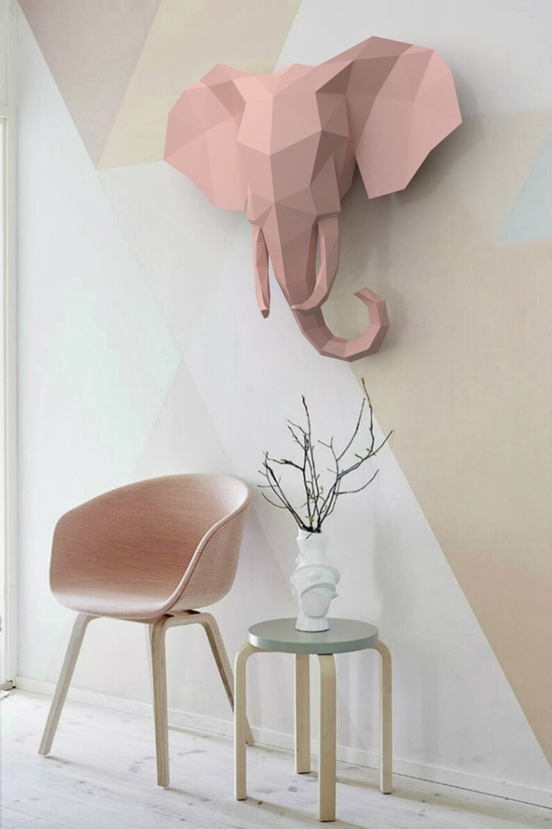 May include: A pink origami elephant head sculpture mounted on a white wall with a pink and white geometric pattern. A pink chair and a small wooden stool with a light green top are in the foreground.