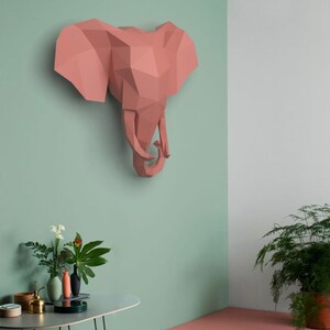 May include: A pink origami elephant head sculpture mounted on a light green wall. The sculpture is made of paper and has a geometric design. The wall is painted in a light green color. There is a table with plants in the foreground.