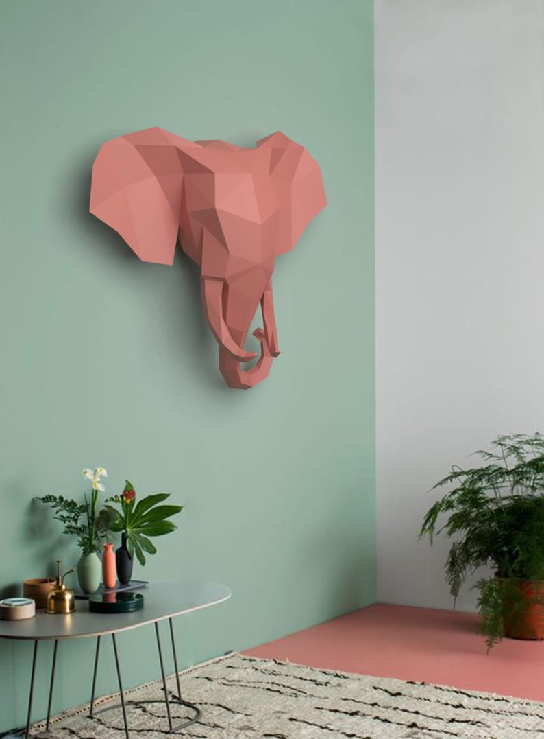 May include: A pink origami elephant head sculpture mounted on a light green wall. The sculpture is made of paper and has a geometric design. The wall is painted in a light green color. There is a table with plants in the foreground.