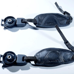 May include: Two black leather camera wrist straps with adjustable nylon straps and quick-release connectors. The straps have a padded design for comfort and feature a metal attachment point. The brand name is visible on the leather.