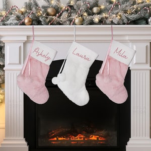May include: Three pink and white Christmas stockings with pom-poms hanging on a white fireplace mantel. The stockings are personalized with the names "Ashley", "Laura", and "Mila" in pink script.