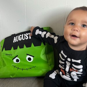 May include: A green Halloween trick-or-treat bag with a Frankenstein face and the name "AUGUST" printed on it. A baby wearing a black skeleton costume is holding the bag.
