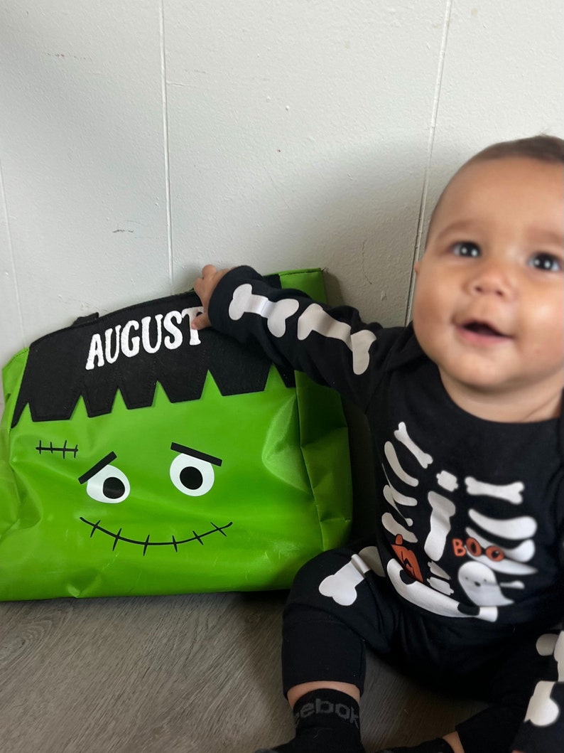 May include: A green Halloween trick-or-treat bag with a Frankenstein face and the name "AUGUST" printed on it. A baby wearing a black skeleton costume is holding the bag.