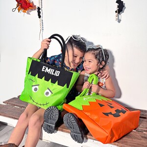 May include: Two children, one holding a green Frankenstein-themed trick-or-treat bag with the name "Emilio" on it, and the other holding an orange pumpkin-themed trick-or-treat bag with the name "Amelia" on it.