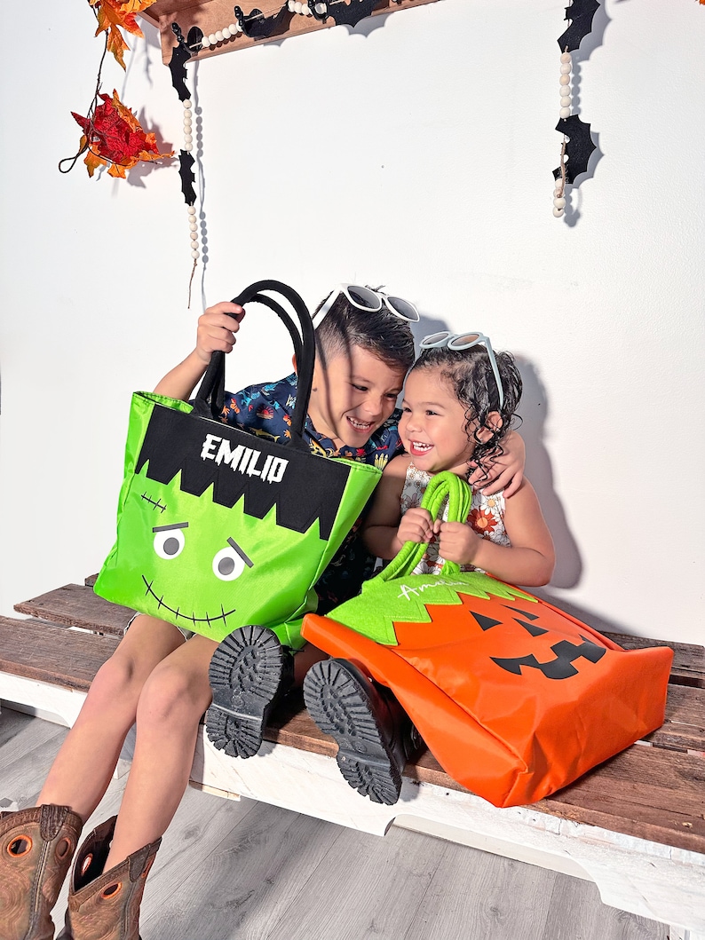 May include: Two children, one holding a green Frankenstein-themed trick-or-treat bag with the name "Emilio" on it, and the other holding an orange pumpkin-themed trick-or-treat bag with the name "Amelia" on it.