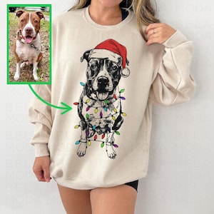 Op de afbeelding: Een beige sweater met ronde hals voorzien van een grafische illustratie van een hond met een rode kerstmuts, omwikkeld met kleurrijke kerstverlichting. Het ontwerp is gebaseerd op een foto van een bruin-witte hond in de linkerbovenhoek.