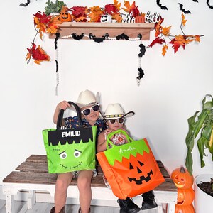 May include: Two children wearing cowboy hats and sunglasses hold personalized Halloween trick-or-treat bags. The boy's bag is green with a Frankenstein face and the text "EMILIO". The girl's bag is orange with a jack-o'-lantern face and the text "Amelia".