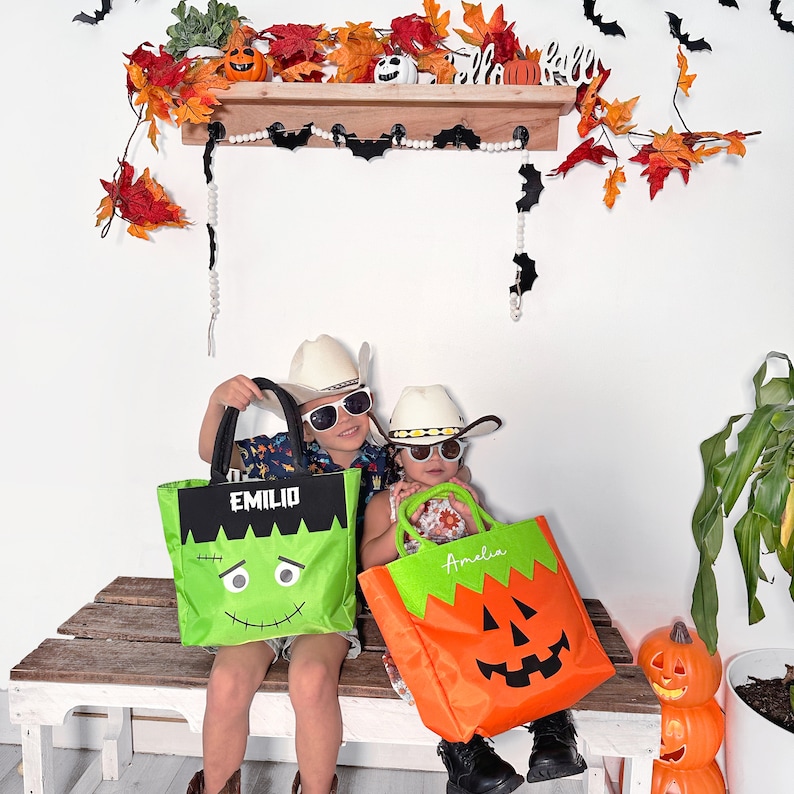 May include: Two children wearing cowboy hats and sunglasses hold personalized Halloween trick-or-treat bags. The boy's bag is green with a Frankenstein face and the text "EMILIO". The girl's bag is orange with a jack-o'-lantern face and the text "Amelia".