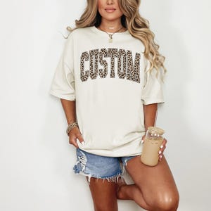 May include: Cream-colored t-shirt with the word "CUSTOM" in a leopard print design. The shirt has short sleeves and a relaxed fit. The model is wearing denim shorts and holding a glass of iced coffee.