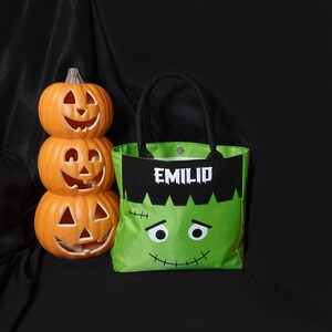 May include: Green and black Halloween trick-or-treat bag with a Frankenstein face and the name "Emilio" embroidered on the front. The bag is next to a stack of three orange jack-o'-lantern pumpkins.