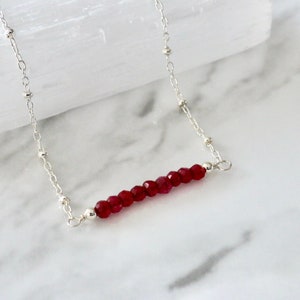 Op de afbeelding: Een zilveren ketting met een staaf van rode edelsteen kralen. De ketting ligt op een wit marmeren oppervlak.