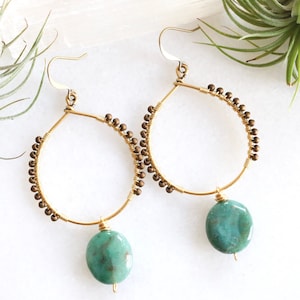 Puede incluir: Pendientes de aro dorados con un colgante de turquesa. Los aros están decorados con pequeñas cuentas doradas.