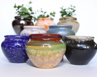 May include: A collection of eight small ceramic pots in various colors and patterns, including blue, green, brown, black, and white. The pots are all different shapes and sizes, and some have a glossy finish.