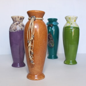 May include: Four ceramic vases in different colors and styles. The vase in the center is brown with a textured surface and a decorative rope and bead accent. The vase to the left is purple with white speckles. The vase to the right is green with white speckles. The vase in the back is teal with a decorative shell accent.