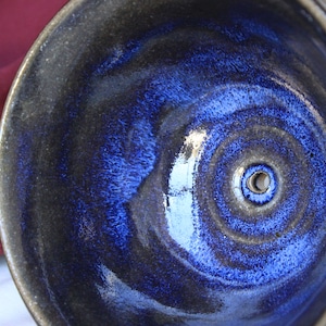 May include: A dark blue ceramic bowl with a textured glaze and a small hole in the center. The bowl has a deep, round shape and a slightly raised rim.